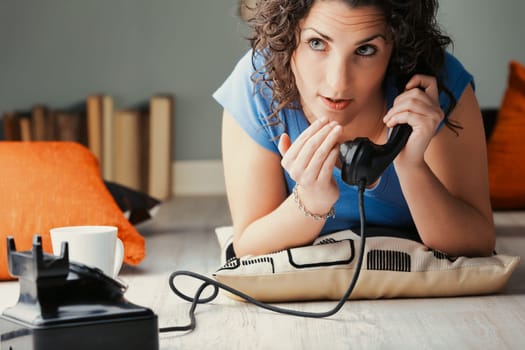 Lounging on the floor amidst cushions, a woman excitedly whispers into a vintage black phone. Amidst the pleasure of gossip, she's oblivious to the fact she'll be discussed next in the rumor mill