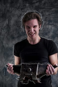 On a dramatic grunge-black backdrop, a young man in a black shirt lifts an anvil with notable effort, yet succeeds. His robust muscles aid him. Even strained, joy emerges through his laughter