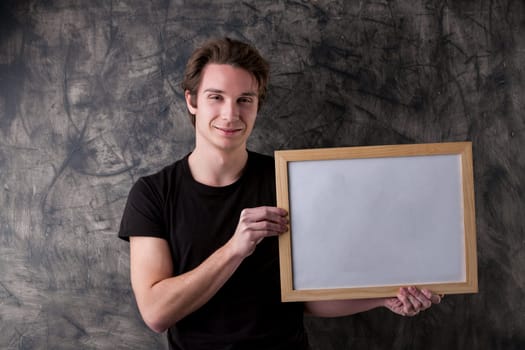 Student or professor, a young man holds a framed white copyspace sign. Behind him is a wiped blackboard. His reassuring smile invites you to learn, hinting at the benefits of education