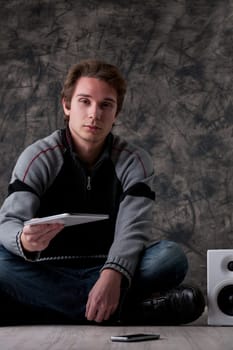 Young man in a grey and black sweater stands indoors, near white hi-fi speakers and a smartphone. He extends a digital tablet, promoting mobile tech, apps, and online tools for work and entertainment