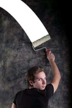 On a black grunge-marked board, a dashing youth with light-brown hair in a black tee uses a roller to craft a perfect white section intended for COPYSPACE