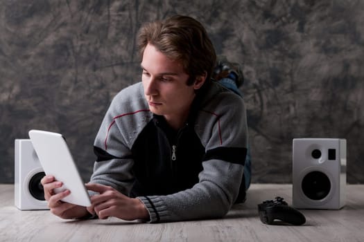 Young man lies on the floor with a white digital tablet, gaming controller, and speakers nearby. Digital entertainment is great if you don't have time to go out. Content is vast, but ownership is fadi