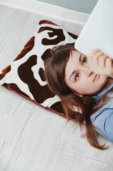 Overhead view: young woman's face on leopard-patterned pillow. Lying on a home floor, she enjoys her white digital tablet for games, apps, and web exploration in a secure middle-class setting