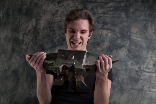 Against a stark grunge scene, a young male raises an anvil with visible strain. His muscular physique is evident. Amidst exertion, a chuckle escapes