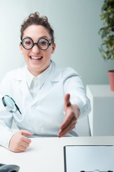 Across a desk, a nerdy-appearing woman in a white coat presents her hand. The subconscious pull of beauty, as identified by psychology, serves as an unchosen but real advantage, especially during job