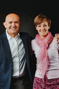 In a dimly lit studio, a long-married elder couple beams. His baldness is accentuated by a blue jacket and pale blue shirt her attire, predominantly pink, subtly echoes the red tints in her hair