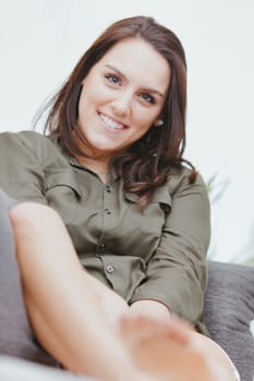 A close-up portrait of a young woman with a big smile and long brown hair sitting on her white couch at home. She's happy and relaxed, wearing short shorts and a military green shirt. Her legs are smo