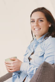 A happy and smiling woman sits on her white couch at home, enjoying her free time with style. She sips a hot drink from a green mug while wearing a blue denim shirt and skirt. Sunlight streams in thro