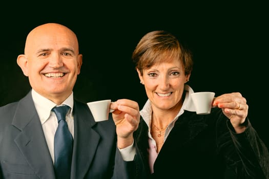 Portrayed elegantly against black, two wealthy elderly enjoy Italian espresso on their European voyage. While he's bald with jacket and shirt, she flaunts boyish red hair. Both toast to the health of