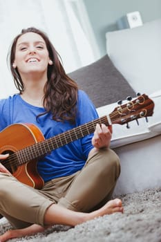 A girl, a young woman, plays the guitar with crossed legs on the carpeted sofa in her home. The acoustic guitar amuses her, and she loves art as well as music. Creativity will disappear, replaced by a