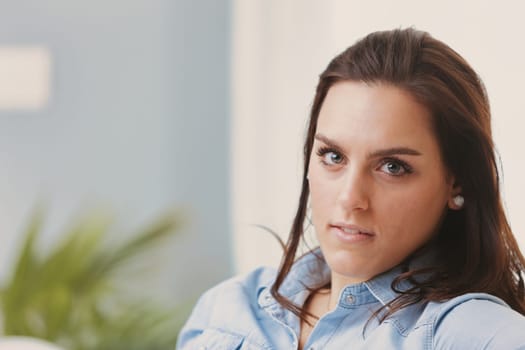 A close-up portrait of a young woman with brown hair and light-colored eyes, wearing a denim shirt and indoors. In the blurred background, a plant and a sunlit window are visible. Her intelligent gaze