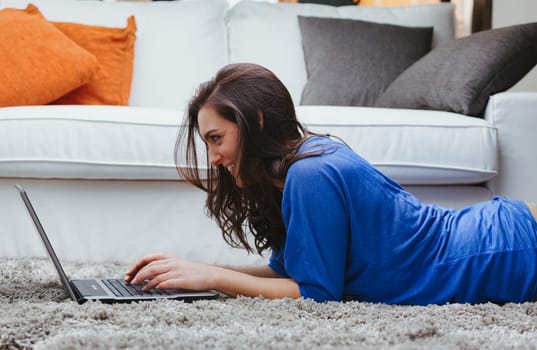 A young woman on the soft and comfortable carpet of her home uses a laptop computer. Is she working, studying, or having fun? Thanks to the wifi and broadband that quickly reach the internet, she does