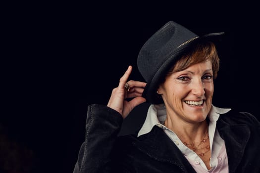 Against a black background, wearing a Borsalino-style hat and a dark jacket, a lady laughs as she does the moves of a 1950s Hollywood dancer.