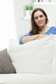A proud and satisfied woman, confident, happy, leaning against the back cushion of her home sofa. The decor is bright, and the sunlight filters through the bright curtains from the window. She is beau