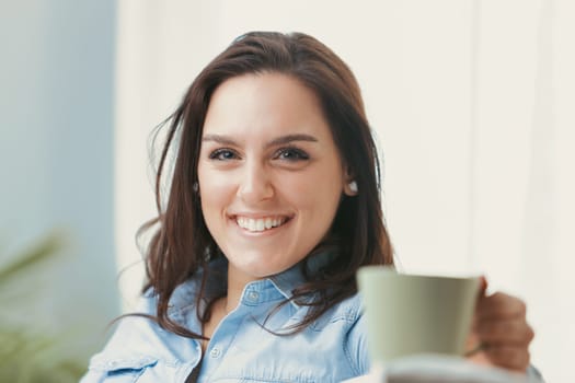 A happy and smiling woman sits on her white couch at home, enjoying her free time with style. She sips a hot drink from a green mug while wearing a blue denim shirt and skirt. Sunlight streams in thro