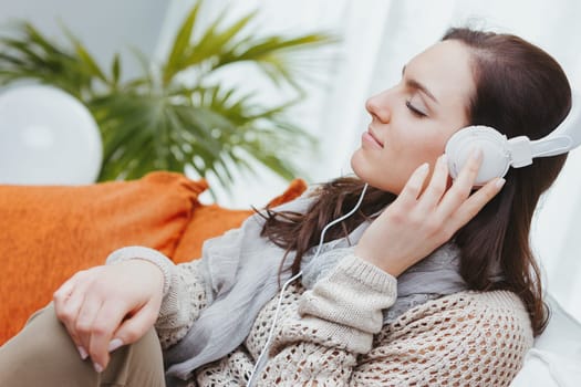 A woman dressed in shades of brown is sitting barefoot on her home sofa. She's listening to music with white headphones, or maybe to the radio or an interesting podcast. Relaxing and entertaining at h
