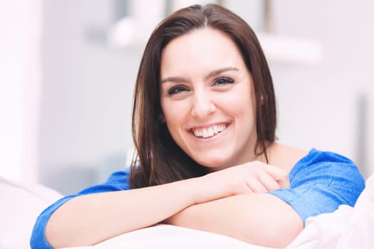 A young, happy, and dreamy woman smiles with her head resting on her crossed arms, which are in turn leaning against the cushion of the sofa. She imagines, invents, and reasons. She imagines a way to