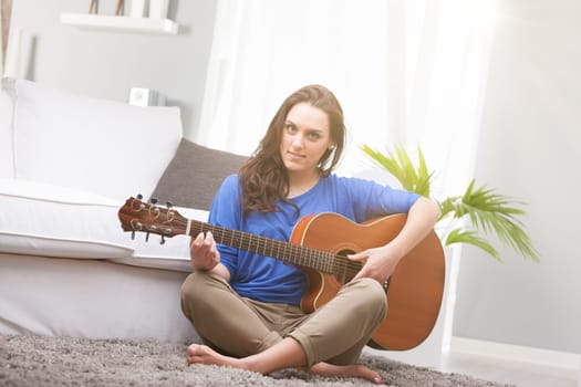 A girl, a young woman, plays the guitar with crossed legs on the carpeted sofa in her home. The acoustic guitar amuses her, and she loves art as well as music. Creativity will disappear, replaced by a