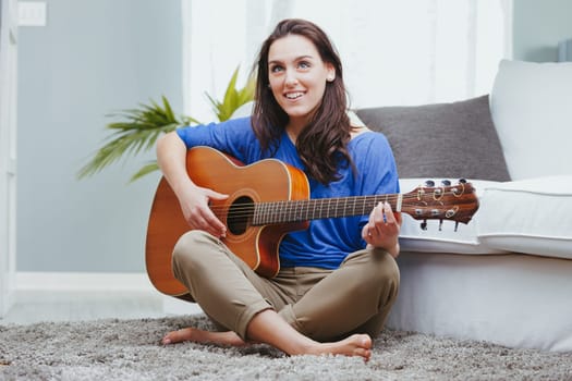 A girl, a young woman, plays the guitar with crossed legs on the carpeted sofa in her home. The acoustic guitar amuses her, and she loves art as well as music. Creativity will disappear, replaced by a