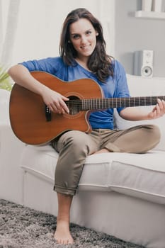 A girl, a young woman, plays the guitar with crossed legs on the carpeted sofa in her home. The acoustic guitar amuses her, and she loves art as well as music. Creativity will disappear, replaced by a