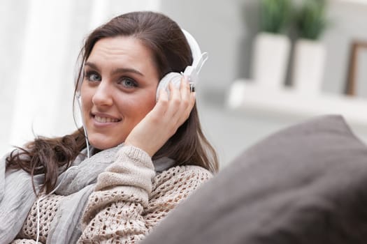 A woman dressed in shades of brown is sitting barefoot on her home sofa. She's listening to music with white headphones, or maybe to the radio or an interesting podcast. Relaxing and entertaining at h