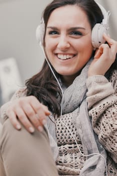 A woman dressed in shades of brown is sitting barefoot on her home sofa. She's listening to music with white headphones, or maybe to the radio or an interesting podcast. Relaxing and entertaining at h