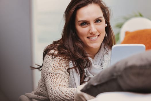 Portrait of a young woman with beautiful eyes that convey intelligence, holding a white digital tablet. She sits on the carpet at the foot of the sofa, enjoying culture and entertainment from the inte