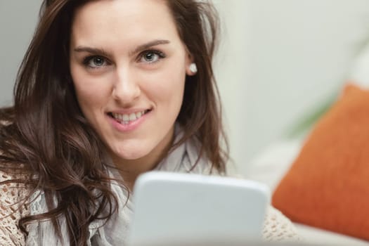 Portrait of a young woman with beautiful eyes that convey intelligence, holding a white digital tablet. She sits on the carpet at the foot of the sofa, enjoying culture and entertainment from the inte