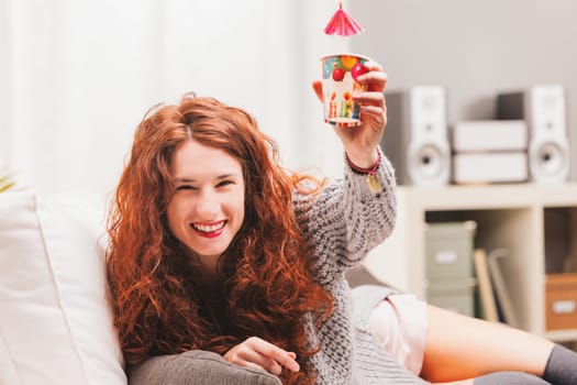 Embodying a carefree lifestyle, a radiant redhead on a white sofa revels in festivities. In a mix of intimate apparel and a woolen top, she beams with joy, raising celebratory items in cheer