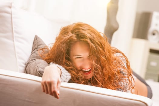Woman, joyously laughing, sprawled on a sofa, represents quality time: relaxation, happiness, carefreeness. Long, red-haired young lady in her living room