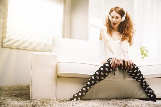 In her bright home, a young, slender woman with striking red hair loses herself in music. Dressed casually, she sits on a white couch, indulging in the rhythms from her white headphones