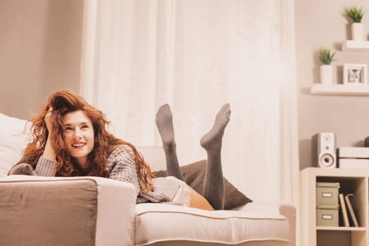 Dreaming of weekends or vacations, possible dates or career progress, a woman lays relaxed in her intimate wear, sweater, and knee-high stockings on her couch. Her joy and satisfaction are evident, co