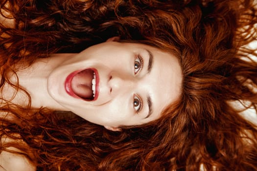 Overhead shot of a spirited young woman with wide-open mouth showcasing flawless teeth. Her vibrant eyes and red lipstick juxtapose against her long red hair, exuding joy and magnetism