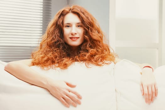 Peeking from behind plush sofa cushions, a joyous woman beams amid her fiery long, billowy red hair. Lounging gracefully on her white couch, her slender form is evident