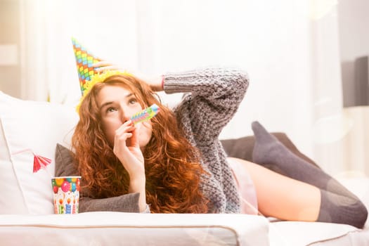 On her white couch, a slender woman with flowing red hair combines comfort with celebration. Dressed in intimates and a soft wool sweater, she laughs and smiles, embodying a festive lifestyle