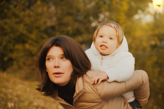 Enjoying the outdoors, a blonde child climbs atop her lovely mother's shoulders. They're both snug against autumn's chill, sharing a day of boundless love and company