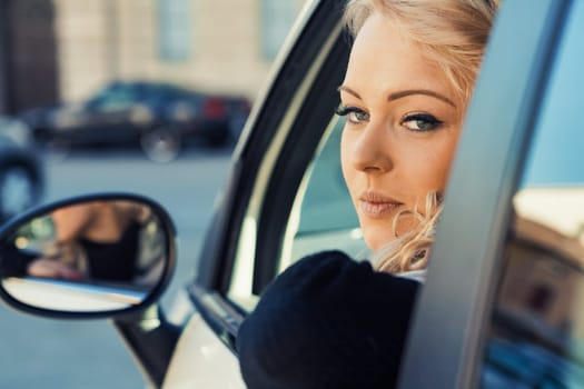 A young woman with blonde curls, impeccable makeup, and manicure embarks on a European adventure in a fashionable car. Her enchanting eyes sparkle as she smiles, shielding herself from sunlight