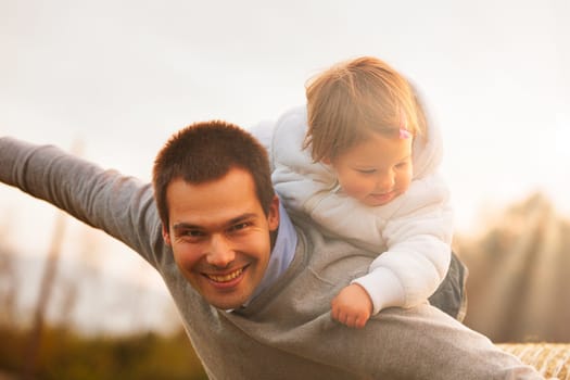 Laughter echoes in the field as a father and daughter engage in a playful game of pretend flight at dusk