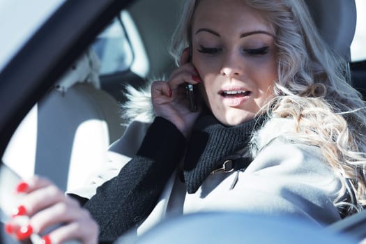 Beautiful young blonde woman with long hair, very outgoing, uses her phone and smartphone while driving a fashionable small car in Europe, about to have fun with friends or as an independent tourist,