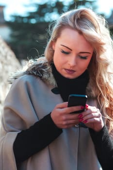 Young woman with long, curly, perfectly permed blonde hair, perfect makeup and manicure, is a tourist in a European city with medieval villages. Using her phone to talk, chat, or access Wi-Fi for hist