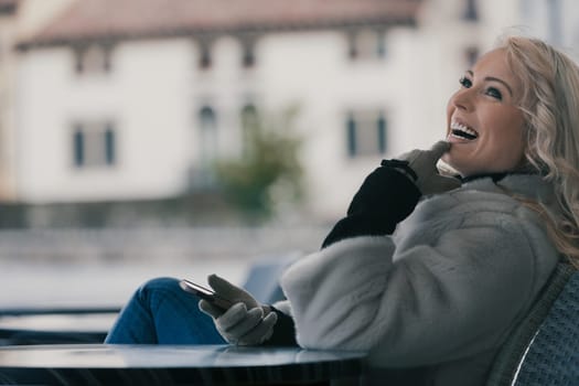 Beautiful young blonde woman sitting at a bar, having fun, laughing at something hilarious on her smartphone or a received message. She's a tourist in a European city, enjoying the fresh air, dressed