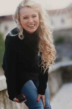 Young woman with long, curly, perfectly permed blonde hair, perfect makeup and manicure, is a tourist in a European city with medieval villages. Using her phone to talk, chat, or access Wi-Fi for hist