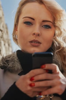 Young woman with long, curly, perfectly permed blonde hair, perfect makeup and manicure, is a tourist in a European city with medieval villages. Using her phone to talk, chat, or access Wi-Fi for hist
