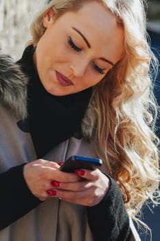 Young woman with long, curly, perfectly permed blonde hair, perfect makeup and manicure, is a tourist in a European city with medieval villages. Using her phone to talk, chat, or access Wi-Fi for hist