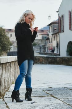 Young woman with long, curly, perfectly permed blonde hair, perfect makeup and manicure, is a tourist in a European city with medieval villages. Using her phone to talk, chat, or access Wi-Fi for hist