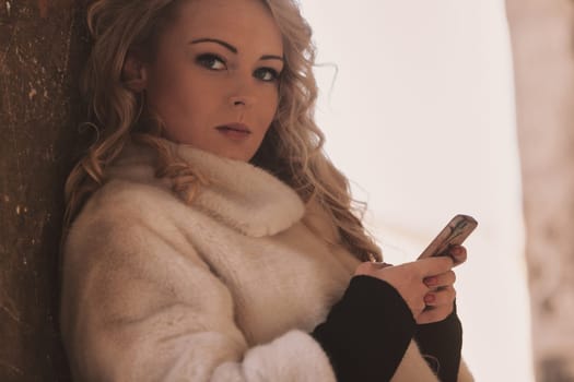 Young woman with long, curly, perfectly permed blonde hair, perfect makeup and manicure, is a tourist in a European city with medieval villages. Using her phone to talk, chat, or access Wi-Fi for hist