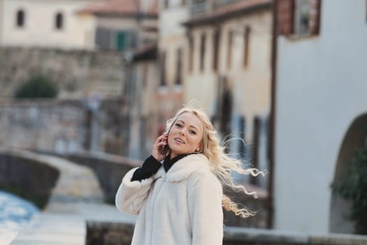 Young woman with long, curly, perfectly permed blonde hair, perfect makeup and manicure, is a tourist in a European city with medieval villages. Using her phone to talk, chat, or access Wi-Fi for hist