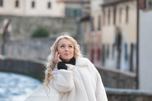 Young woman with long, curly, perfectly permed blonde hair, perfect makeup and manicure, is a tourist in a European city with medieval villages. Using her phone to talk, chat, or access Wi-Fi for hist