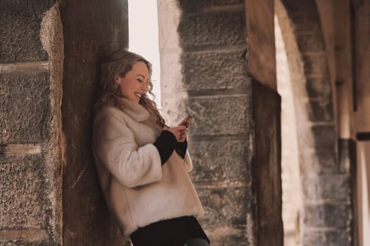 Young woman with long, curly, perfectly permed blonde hair, perfect makeup and manicure, is a tourist in a European city with medieval villages. Using her phone to talk, chat, or access Wi-Fi for hist