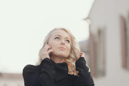 Young woman with long, curly, perfectly permed blonde hair, perfect makeup and manicure, is a tourist in a European city with medieval villages. Using her phone to talk, chat, or access Wi-Fi for hist